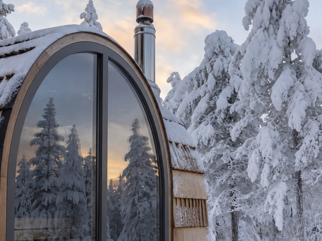 Ośnieżona sauna z łukowym oknem odbijającym zimowy las.